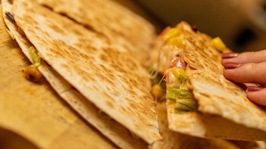 Eine Hand nimmt ein Viertel eines runden Quesadillas. Darin ist geschmolzener Käse und Gemüse.