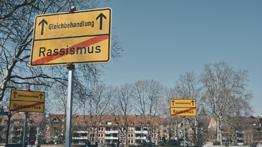 Dreimal dieselben gelben Orteingangsschilder auf einem Platz. Darauf ist Rassismus durchgestrichen und Gleichbehandlung mit einem Pfeil gekennzeichnet