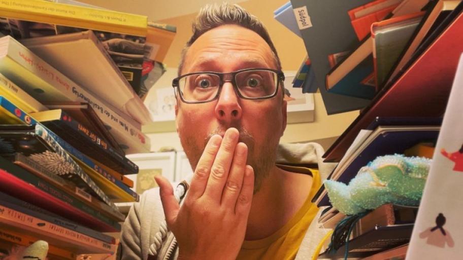 Ein mann mit einer brilole ist zu sehen zwischen Stapeln von Büchern. Er hält sich die rechte Hand vor den mund. 