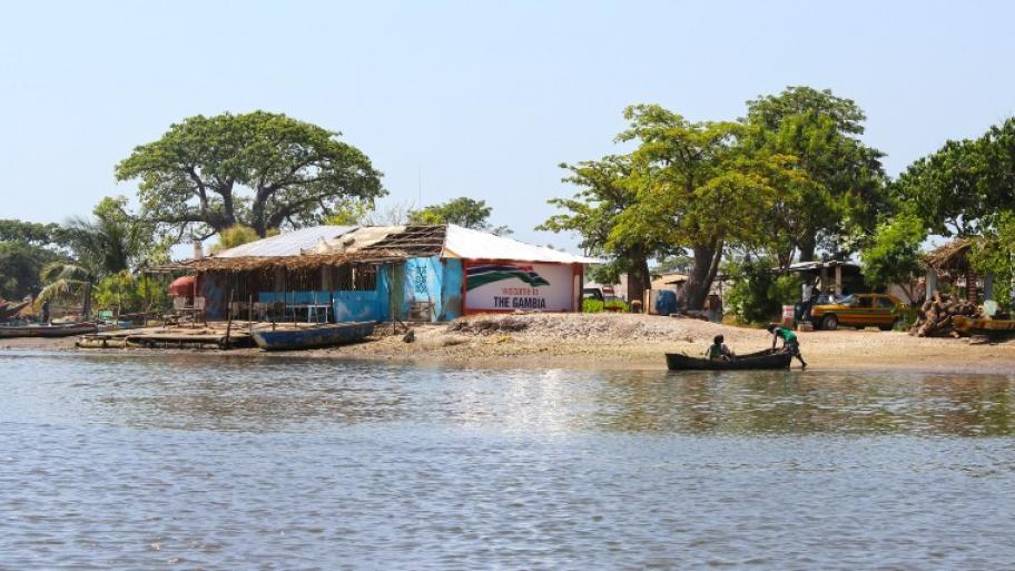 Flussszene in Gambia: vorne der Fluss, dahinter am Ufer ein blaues Haus mit Strohdach, zwei Menschen in einem kleinen Boot, im Hintergrund weitere Hütten, Bäume, ein Auto