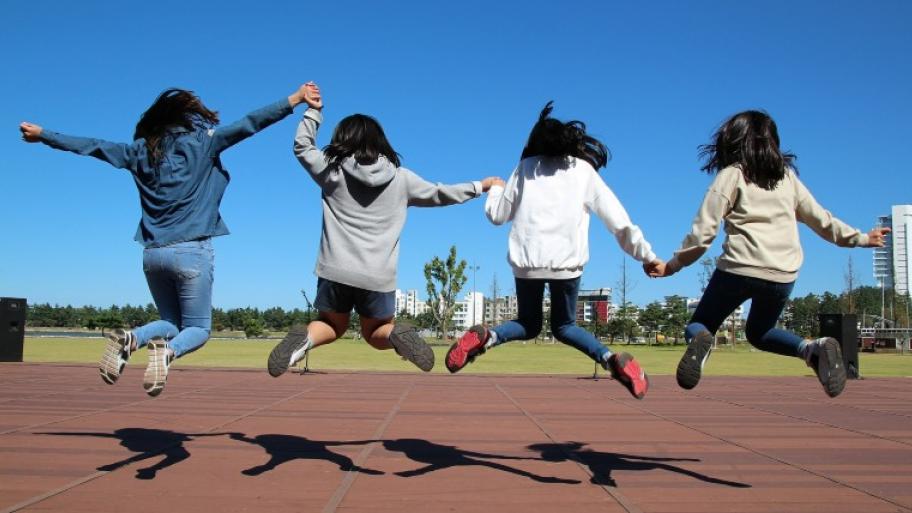 4 Kinder von hinten auf roten Steinplatten: sie halten sich an den Händen und springen gemeinsam in die Luft, die Beine sind angewinkelt, die schwarzen Haare fliegen, im Hintergrund eine grüne Wiese und Häuser