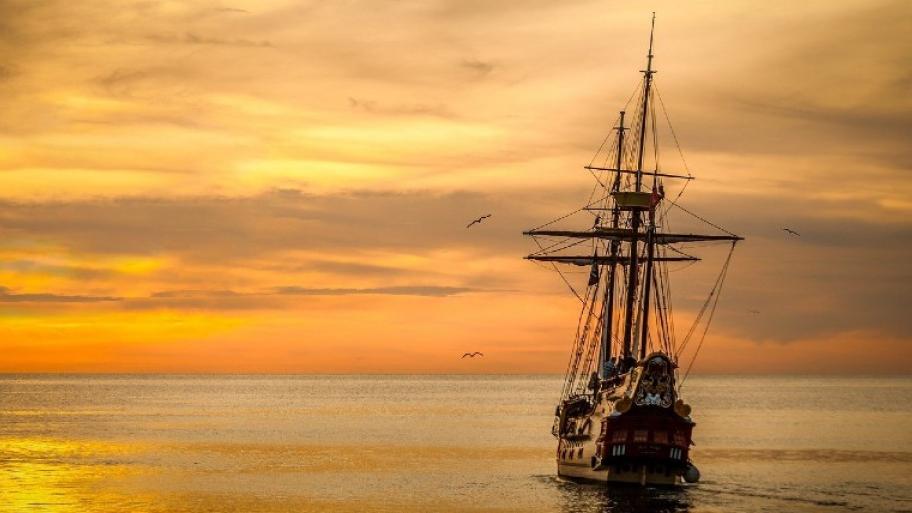 Piratensegelschiff von hinten auf der rechten Seite des Bildes segelt auf einen Sonnenuntergang zu.