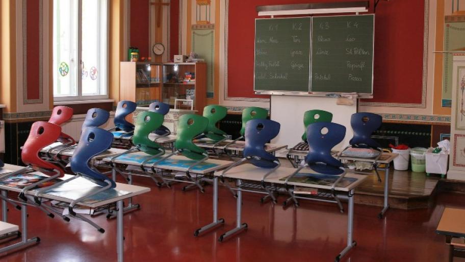 ein Klassenzimmer mit rotem Fußboden, darin hinten eine große Tafel, links ein gro0es Fenster, drei Reihen mit weißen Tischen, darauf hochgestellt Stühle in rot, blau, und grün