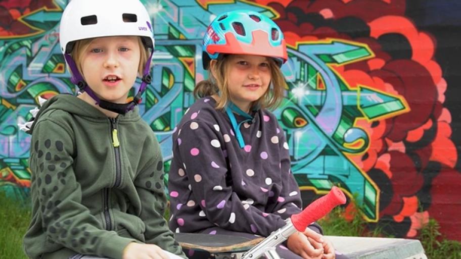 ein Junge und ein Mädchen sitzen auf einer steinernen Bank, beide tragen einen Fahrradhelm und einen bunten Kapuzenpullover, im Hintergrund eine bunte Graffiti-Wand