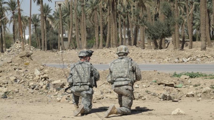 zwei Soldaten von hinten in Uniform knien auf sandigem Boden, dahinter eine Straße, Schuttberge, staubige Palmen