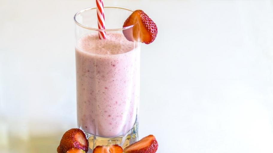 auf weißem Untergrund ein Glas gefüllt mit rötlichem Erdbeer-Smoothie, darin ein rot-weißer Papiertrinkhalm, mehrere Erdbeeren zur Dekoration davor