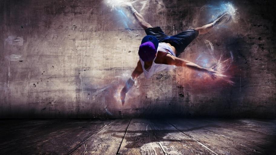 ein Hiphop-Streetdancer macht einen akrobatischen Dancemove und fliegt durch die Luft, von seinen Händen und Füßen gehen bunte Blitze aus, er trägt Jeans, T-Shirt und ein blaues Basecap, Betonwand im Hintergrund