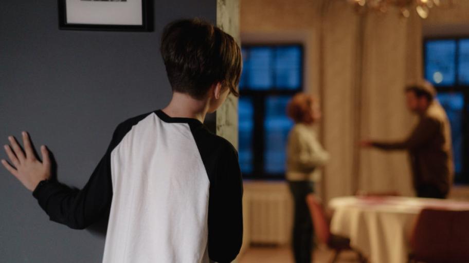 Ein Junge mit braunen Haaren und weißem Tshirt steht an einer grauen Wand und schaut in ein Zimmer, welches hell erleuchtet ist. Man sieht ihn nur von hinten. In dem Zimmer streiten sich ein Mann und eine Frau. Der Teil des Bildes ist verschwommen.