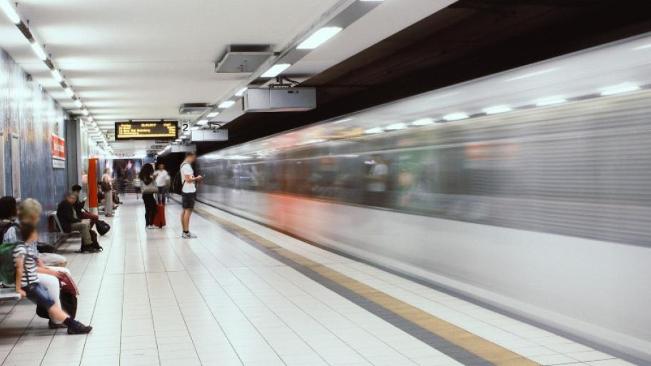 eine silberne U-Bahn fährt in den unterirdischen Bahnhof ein, einige Menschen sitzen oder stehen am Bahnsteig