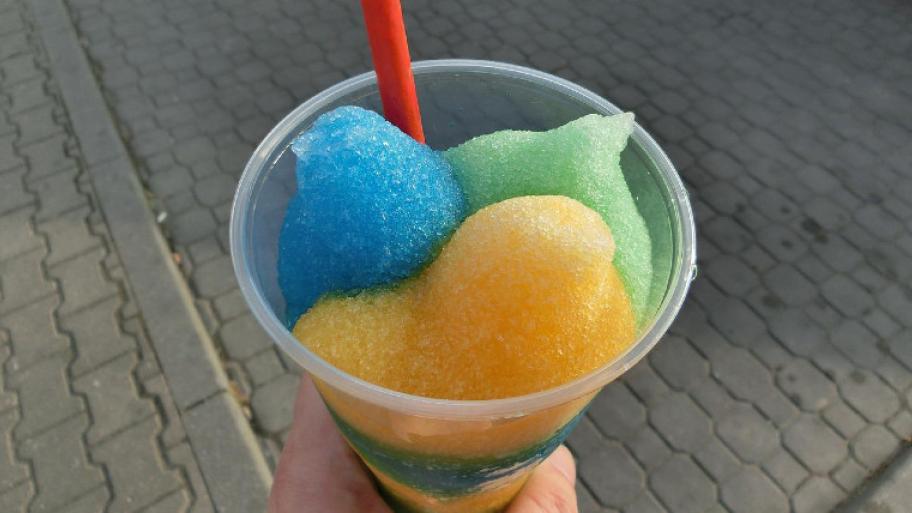 Ein durchsichtiger Plastikbecher ist gefüllt mit einem, blauen, grünen und gelben Wassereisklecks. Oben schaut ein roter Strohhalm heraus.