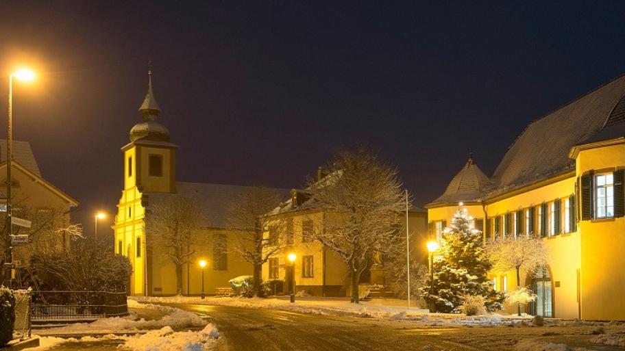 Eine Dorfstraße in der Nacht, die von Laternen beleuchtet wird. Ringsherum stehen Häuser und es liegt Schnee.