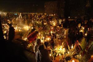 Angehörige feiern auf einem mexikanischen Friedhof den Día de los muertos, es ist dunkel, die Gräber sind mit Blumen dekoriert, es leuchten viele Kerzen