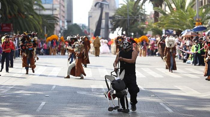 ein Straßenumzug, Menschen mit Masken und bunten Kostümen, mit als Totenkopf geschminkten Gesichtern tanzen auf der Straße, am Rand viele Zuschauer