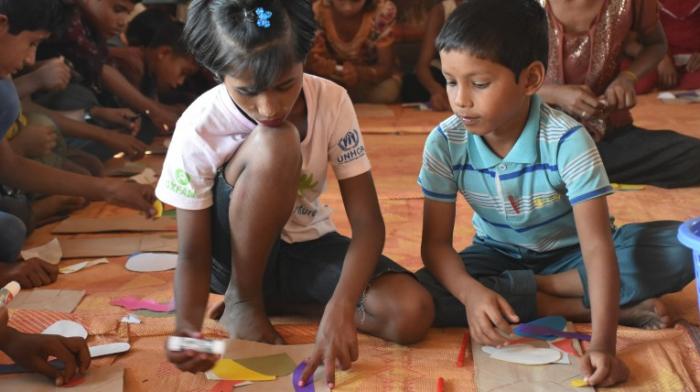 Schulunterricht im Flüchtlingscamp Kutupalong in Bangladesch: zwei Kinder in kurzer Hose und T-Shirt sitzen auf dem Boden eines Raumes und basteln, um sie herum weitere Kinder