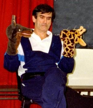 der US-amerikanische Psychologe Marshall B. Rosenberg bei einem Workshop über gewaltfreie Kommunikation 1990, er sitzt auf einem Stuhl und hat über der rechten Hand eine Giraffen-Handpuppe, über der linken eine Wolfs-Handpuppe