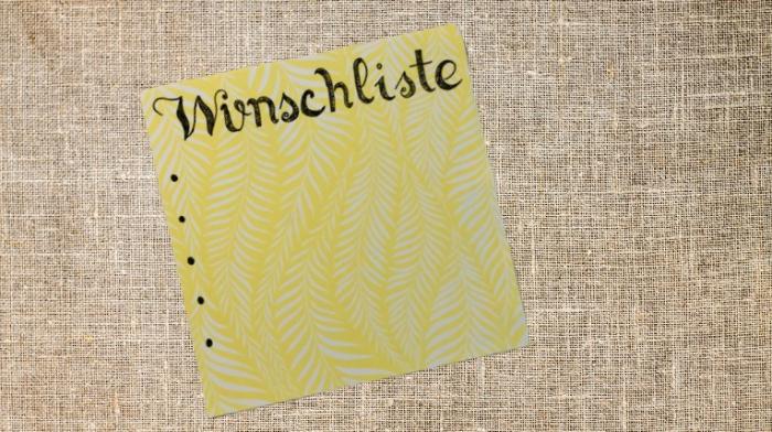 auf einem Textilen, hellen Untergrund liegt schräg ein gelber, quadratischer Zettel, darauf steht in schwarzer Schreibschrift "Wunschliste"