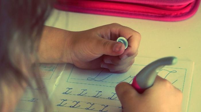 ein Kind von hinten, es schreibt mit einem Füller in der rechten Hand den Buchstaben Z wiederholt in Schreibschrift in ein Schulheft