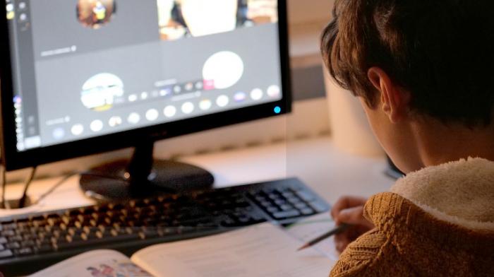 ein Kind sitzt mit Stift in der Hand vor einem Computerbildschirm, darauf läuft ein Online- Klassenmeeting, davor ein geöffnetes Schulbuch