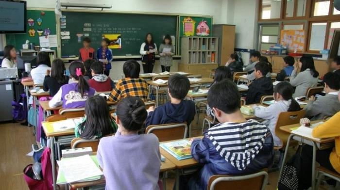 eine Schulklasse sitzt in Reihen im Klassenraum, vier Kinder stehen vorne vor der Tafel und erklären etwas, die Lehrerin sitzt links oben an ihrem Pult