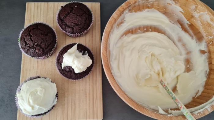 Links stehen vier gebackene Muffins. Auf einem Muffin ist bereits Frosting aufgetragen. Auf einem weiteren ist ein Klecks Frosting drauf. Das Frosting steht rechts.
