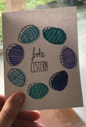 In der Mitte einer Osterkarte steht "frohe Ostern". Die Worte stehen in einem Kranz aus bunten Ostereiern.