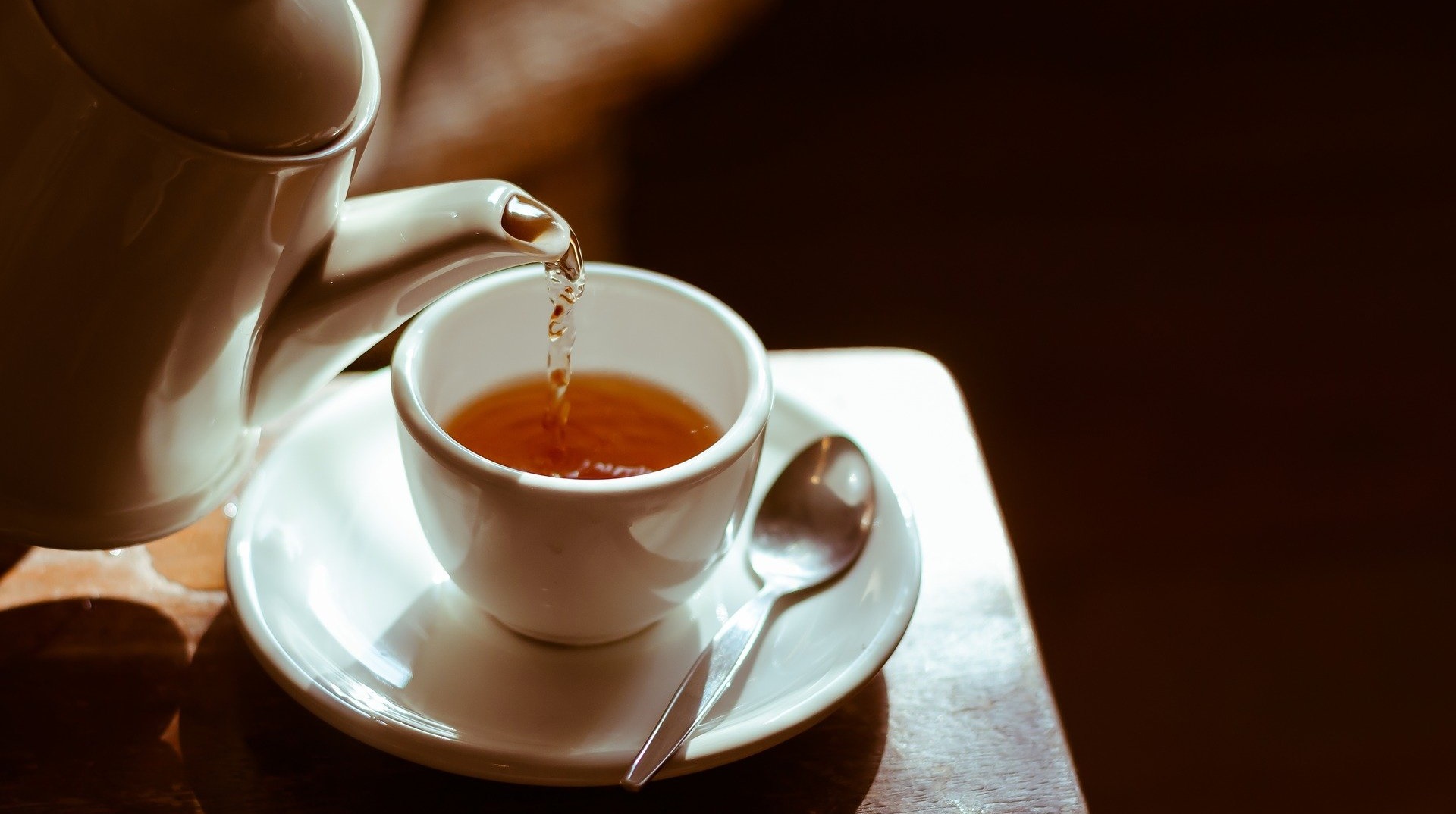 aus einer weißen Teekanne wird bräunlicher Tee in eine weiße Teetasse eingegossen 